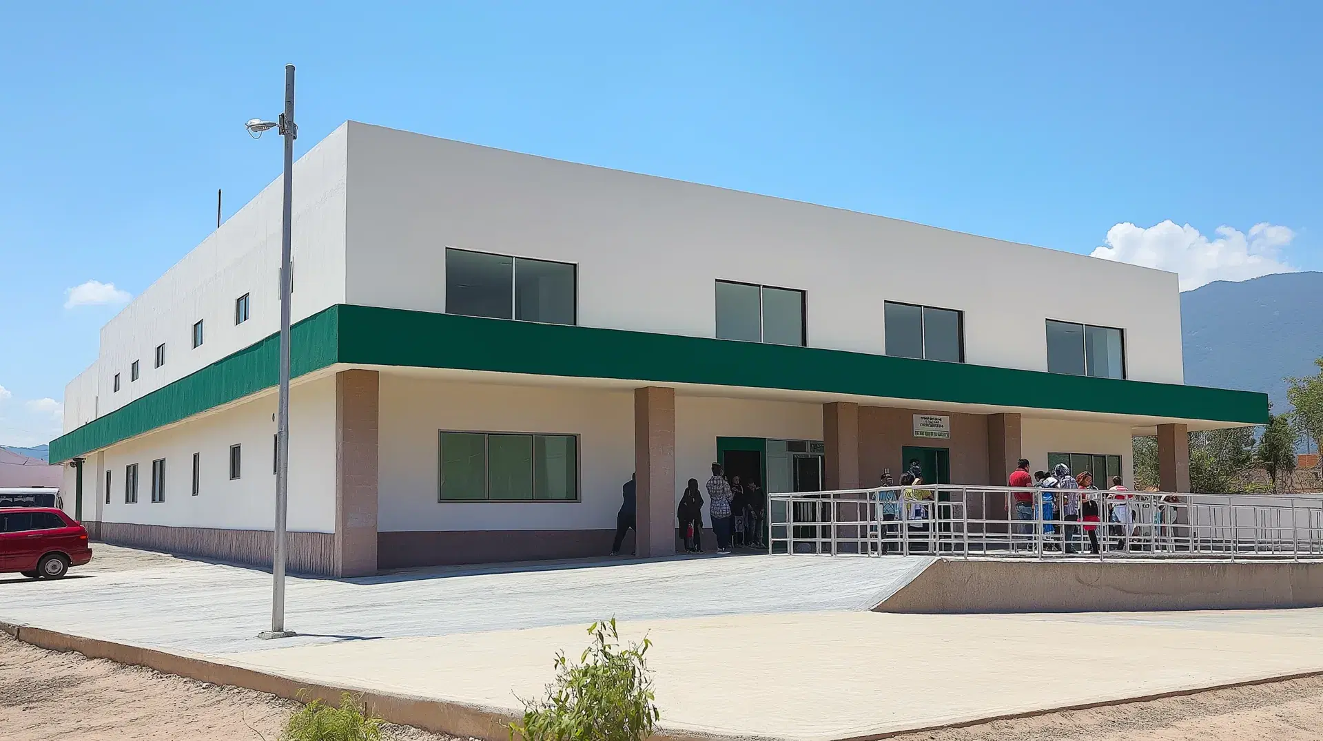 Centro de salud terminado de dos niveles con franja verde en la fachada, rampa de acceso con barandal metálico y personas esperando bajo el pórtico de entrada.